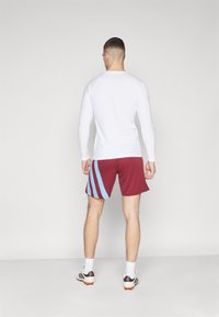 Langsleeve witte sportshirt gecombineerd met bordeauxrode shorts met blauwe diagonale strepen. Model staat van achteren, toont de strakke pasvorm en textuur.