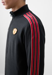 Black zip-up track jacket with high collar, featuring red and white striped accents on sleeves and an embroidered patch on the chest.