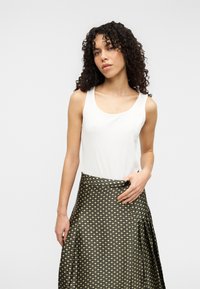 Λευκό αμάνικο τοπ συνδυασμένο με μακριά, σκούρα φούστα με γεωμετρικά σχέδια σε χρυσό και μαύρο. Ομαλό ύφασμα με ελαφριά γυαλάδα.