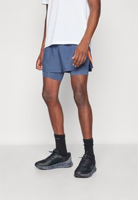Pantalones cortos deportivos navy con detalles en naranja, usados sobre pantalones ajustados interiores. Combinados con zapatillas de correr negras y calcetines negros hasta el tobillo.