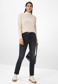 Maglione a collo alto a coste crema abbinato a jeans di denim scuri e sneakers crema. Tiene in mano una borsa nera, in piedi contro uno sfondo chiaro.