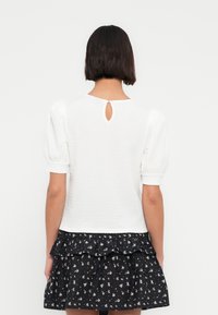 Haut blanc texturé avec manches courtes bouffantes, présentant un détail dos ajouré, associé à une jupe noire à volants imprimée de motifs floraux.