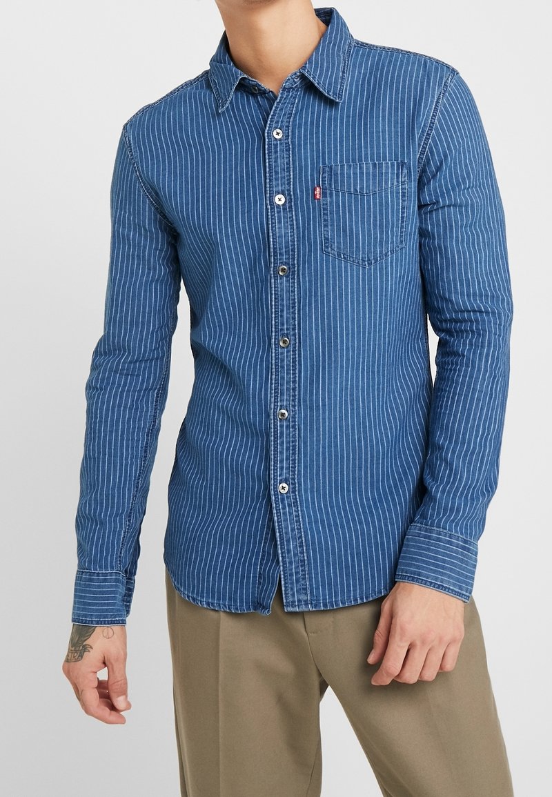 Blauwe denim overhemd met verticale witte strepen, voorzak, knoopsluiting en een klassieke kraag; lange mouwen met knoopmanchen.