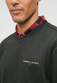 Μαύρη φόρμα με στρογγυλή λαιμόκοψη, διακοσμημένη με χρυσό λογότυπο "TOMMYJEANS" στο στήθος και με κόκκινο και μαύρο καρό πουκάμισο από κάτω.