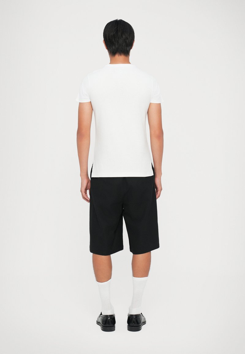 T-shirt blanc à manches courtes, associé à un short noir à longueur genou. Des chaussures noires et des chaussettes blanches complètent la tenue. Design simple avec des couleurs neutres.