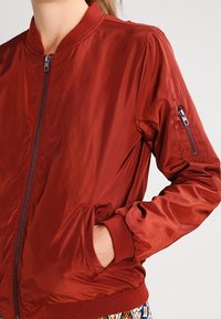 Rote Bomberjacke mit einem Frontreißverschluss, geripptem Kragen und Bündchen, Seitentaschen und einer Reißverschlusstasche am Ärmel. Glattes, glänzendes Material.