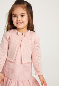 Ljust rosa stickad cardigan med hjärt-mönstrad textur, rund halsringning och hjärtformade knappar. I kombination med en matchande rosa klänning.