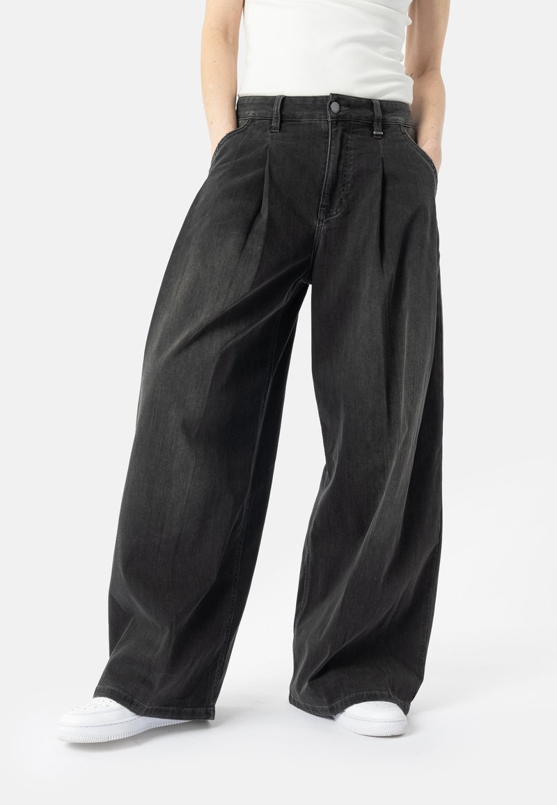 Personne portant un pantalon noir ample à jambes larges, des baskets blanches et un haut blanc sans manches, debout les mains dans les poches.