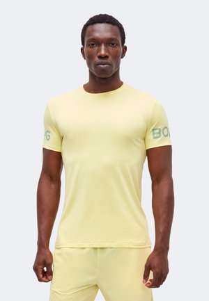 Άνδρας που φορά ανοιχτοκίτρινο πουκάμισο με κοντά μανίκια και αντίστοιχες βερμούδες, στέκεται με χαλαρά τα χέρια σε απλό φόντο.