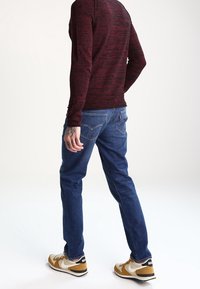 Camisa de manga larga color burdeos, jeans azules y zapatillas beige. Características notables incluyen un tejido texturizado, corte recto y un logotipo en el talón de la zapatilla.