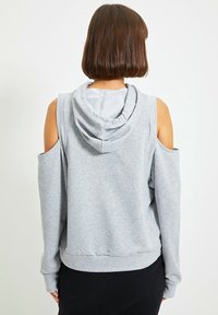 Šedá mikina s odkrytými rameny, stahovací kapucí a žebrovanými manžetami. Materiál se zdá být jemný a strukturovaný, ideální pro každodenní nošení.