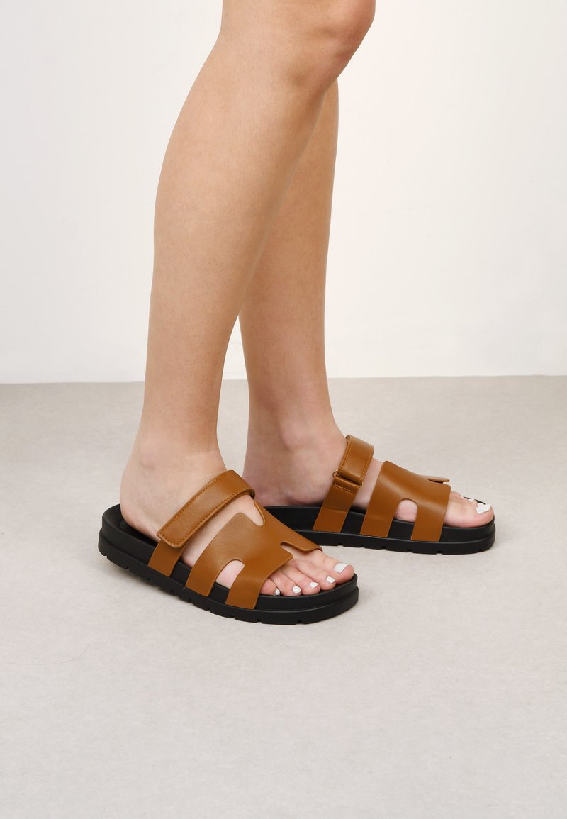 Sandali estivi in pelle marrone, a taglio, con una fascia larga e suola in gomma nera. Presentano una texture liscia e un design minimalista.