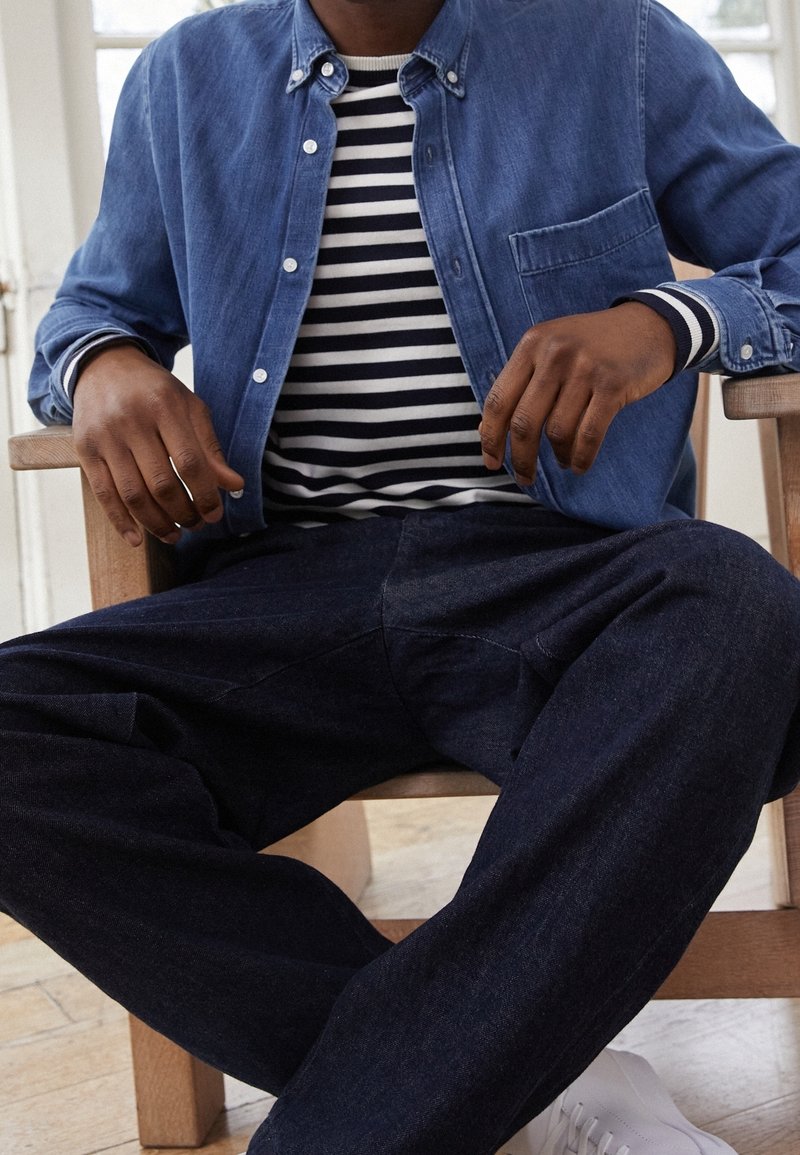 Ljusblå denimskjorta med knappar över en marinblå och vit randig långärmad skjorta, kombinerad med mörka byxor och vita sneakers.