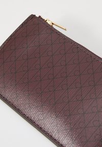 Burgundy kožna novčanica s teksturiranom površinom, s geometrijskim uzorkom crnih trokuta i krugova, naglašena zlatnim patentnim zatvaračem.