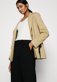 Beige blazer met gestructureerde schouders en revers, gecombineerd met een witte tanktop en zwarte broek. Heeft een losse pasvorm en contrasterende details.