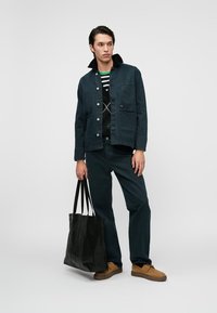 Hombre que lleva chaqueta oscura, pantalones oscuros, suéter con patrón, zapatos beige, sosteniendo una gran bolsa negra, de pie contra un fondo liso.