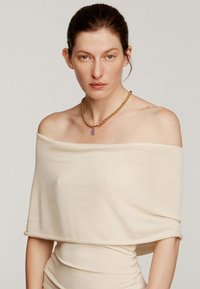 Top claro beige sin mangas, hecho de una tela suave, con un corte relajado. Acompañado de un collar de cadena dorada con un pendant púrpura.