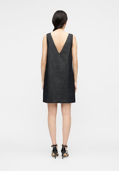 Γυναίκα με μαύρα μαλλιά, φορώντας αμάνικο σκούρο τζιν φόρεμα με βαθύ V στην πλάτη και μαύρα ψηλοτάκουνα σανδάλια, που στέκεται μπροστά σε απλό φωτεινό φόντο.