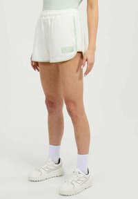 Persona con pantalones cortos elásticos de cintura blanca acanalados con detalles en verde claro, calcetines blancos y zapatillas deportivas blancas, de pie sobre un fondo claro y liso.