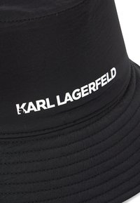Melns buks nierūpīgajā audumā, ar platu malu. "KARL LAGERFELD" logs uz priekšpuses biezām baltajām burtiem.