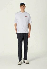 Λευκό κοντομάνικο μπλουζάκι με κόκκινο κείμενο, συνδυασμένο με σκούρους patterned παντελόνες και λευκά αθλητικά παπούτσια με μαύρες λεπτομέρειες.