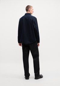 Camisa de algodão azul-marinho com colarinho botão, vista de costas, combinada com calças texturizadas escuras e calçado preto.