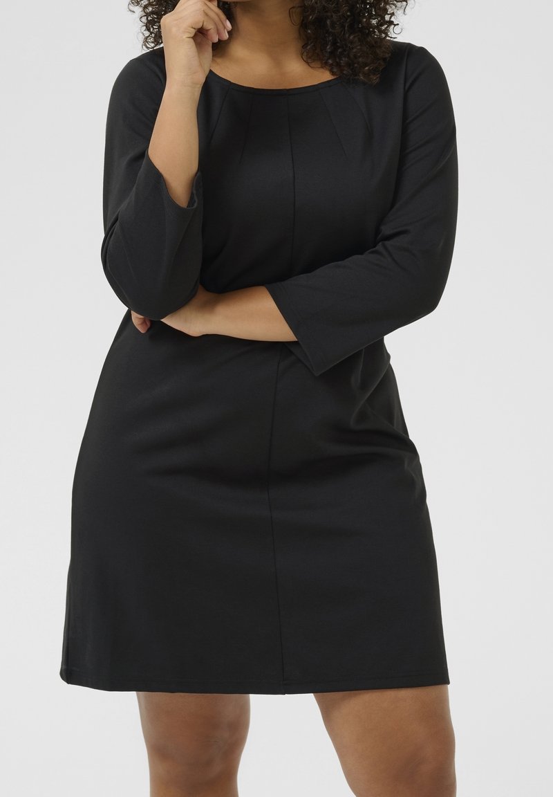Femme aux cheveux bouclés portant une robe noire à manches longues, debout avec un bras croisé et l'autre main près de la bouche.