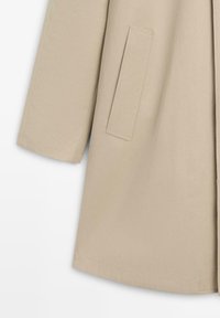 Beige mouw en zijkant van een jas met een verticale zak met net afgewerkte stiknaden op gladde stof.