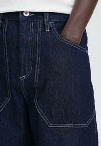 Donkerblauwe spijkerbroek met witte stiksel, voorzien van grote zakken, een brede tailleband en een losse pasvorm. Gestructureerde stof met zichtbare weef.