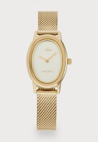 Montre ovale dorée avec bracelet en maille, lunette texturée, cadran blanc minimaliste, aiguilles dorées, logo Lacoste et emblème de crocodile à la position de 12 heures.