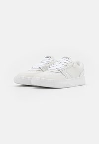 Valged sneakers sünteetilisest nahast, millel on sile tekstuur, lame paelad, perforatsioonidega detailid ja kummist tald mustriga haardega.
