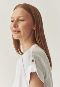 Blusa blanca con mangas remangadas, que presenta acentos de botones de madera. La tela suave y ligera cae con elegancia. Diseño de escote sencillo.