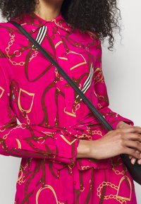 Kvinna iklädd en ljusrosa satin-skjortklänning med ett mönster av bruna och gyllene kedjor, knuten vid midjan, hållande en svart axelremsväska.