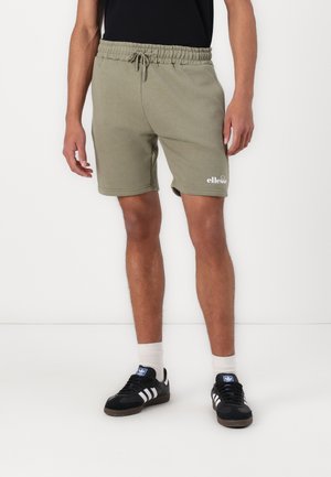Man draagt olijfgroene shorts met trekkoord en zwarte Adidas sneakers met witte strepen, staand tegen een witte achtergrond.