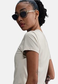 T-shirt in cotone beige con maniche corte, texture liscia. Modello con occhiali da sole trasparenti con lenti scure. Tatuaggio evidente sul braccio.