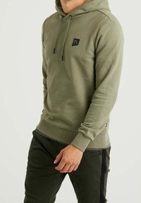 Olivově zelená mikina s kapucí vyrobená z měkké tkaniny, s přední klokaní kapsou a malou černou nášivkou na hrudi. Jednoduchá barva, lehký design.