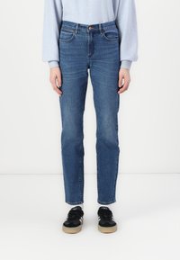 Blå denim jeans med rett ben, fem-lomme design, subtil falming, og en tradisjonell knapplukking. Brukt med svarte sneakers.