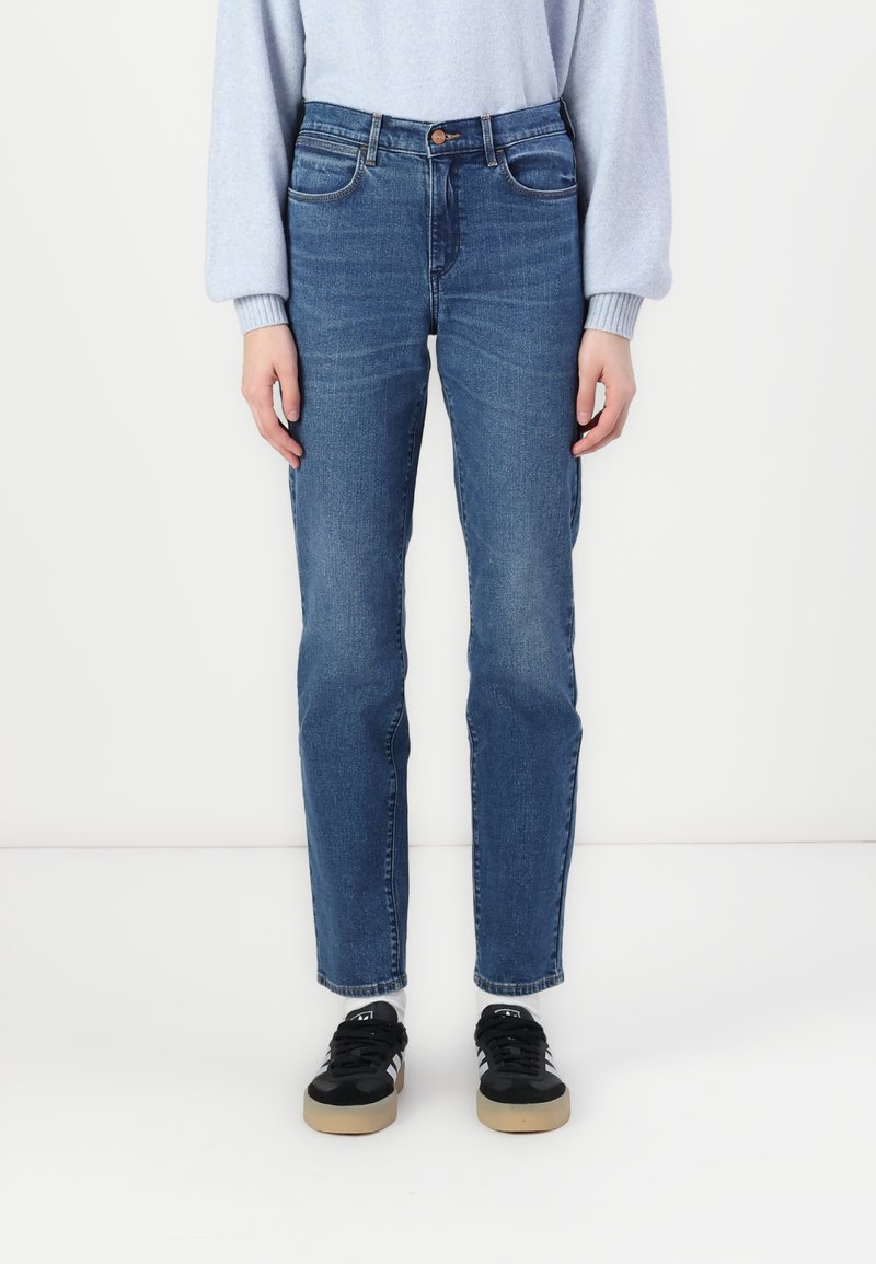 Blå denim jeans med rett ben, fem-lomme design, subtil falming, og en tradisjonell knapplukking. Brukt med svarte sneakers.