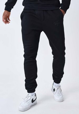 Pantalones de chándal negros con piernas ajustadas, puños elásticos y bolsillos frontales. Presenta un discreto detalle de logo en el costado. Combinados con zapatillas blancas.
