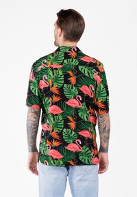 Uomo con le braccia tatuate che indossa una camicia nera a maniche corte decorata con fenicotteri rosa, foglie tropicali verdi e fiori di paradiso arancioni.