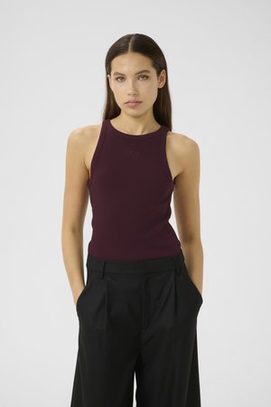 Young woman with long brown hair wearing a sleeveless burgundy top and black wide-leg pants with hands in pockets against a plain backdrop.