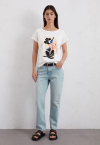 Witte t-shirt met bloemenprint in roze, zwart en blauw, gecombineerd met lichtblauwe denim jeans en zwarte sandalen met gouden accenten.