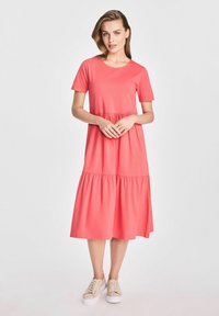 Koralowa bawełniana sukienka z krótkimi rękawami, okrągłym dekoltem i warstwową, rozkloszowaną spódnicą. Noszona z beżowymi sneakersami dla casualowego wyglądu.
