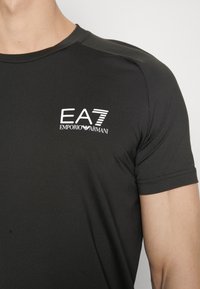 Camiseta atlética negra de manga corta hecha de una tela suave, con un logo blanco de EA7 Emporio Armani en el pecho y un cuello redondo clásico.