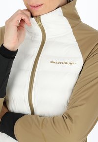 Gefütterte Jacke in Weiß und Beige mit hohem Kragen, Reißverschluss und dem goldenen "SWEDEMOUNT"-Logo. Sie hat kontrastierende schwarze Ärmel.