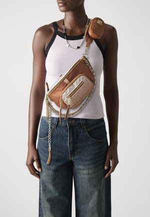 Personne portant un débardeur blanc et un jean bleu avec un sac bandoulière marron doté de plusieurs poches et de détails en chaîne dorée.