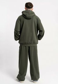 Osoba stoi tyłem do kamery, ubrana w oliwkową bluzę z kapturem i luźne spodnie w tym samym kolorze, widoczne białe trampki na jednolitym tle.