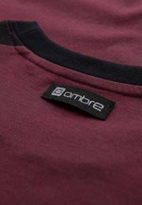 Burgundské tričko s černým žebrovaným výstřihem; obsahuje černou textilní etiketu s bílým textem. Materiál se zdá být hladký a lehký.