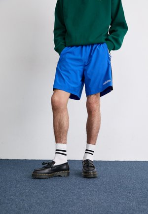 Shorts de sport bleus avec une taille élastique, des accents latéraux noirs et le logo Adidas, associés à des chaussures noires et des chaussettes blanches rayées.