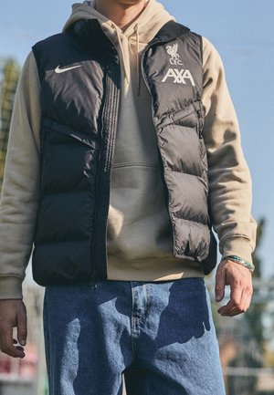 Mężczyzna w beżowej bluzie z kapturem, pod czarną pikowaną kamizelką Nike z logo LFC AXA i niebieskich dżinsach, stojący na zewnątrz na tle czystego nieba.